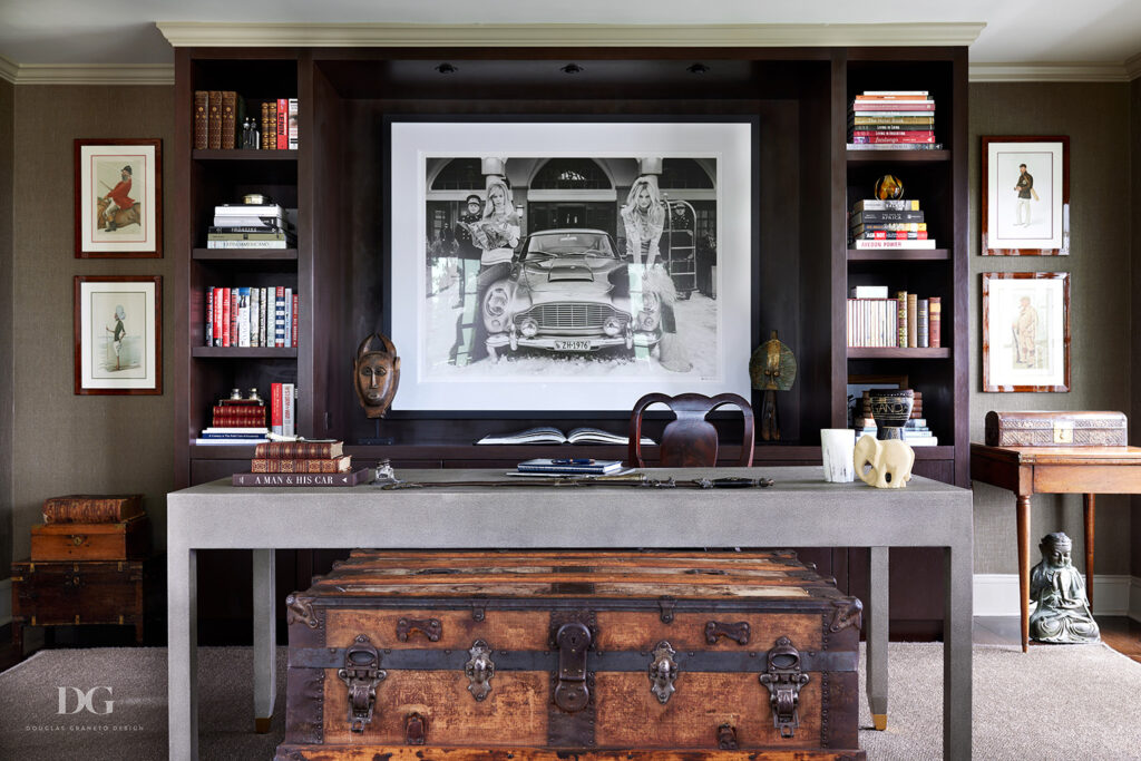 Head on view of a gray desk with dark wood built-in shelving behind. A black and white print directly behind the desk. A vintage chest is in front of it