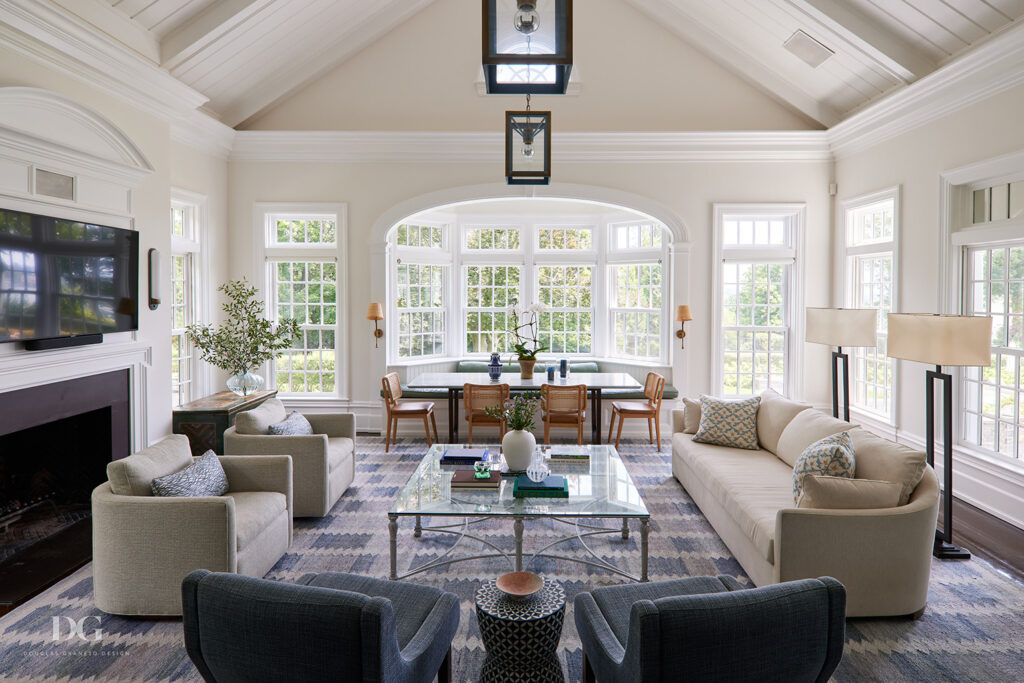 A view of a white family room looking towards a large bay window with built-in window seat. Vaulted ceiling, a pair of blue Espasso armchairs, a Holly Hunt sofa and a blue and white rug
