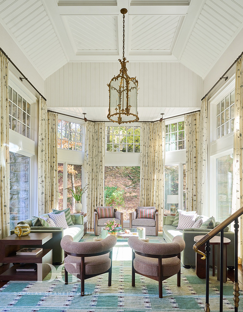 View of a living room with vaulted ceiling, large windows looking out to woodland. A pair of green George Smith sofas and pink Holly Hunt chairs