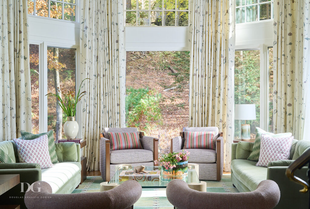 View of a living room with large windows looking out to woodland. A pair of green George Smith sofas and pink Holly Hunt chairs and Chelsea Editions curtains