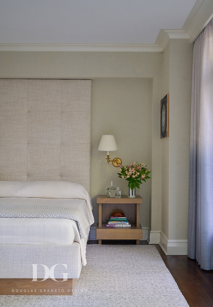 View of half of bed with large custom upholstered headboard and open wood nightstand